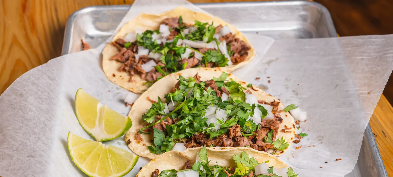 A tray of three street tacos topped with cilantro and onion, with lime wedges alongside, on a wooden table